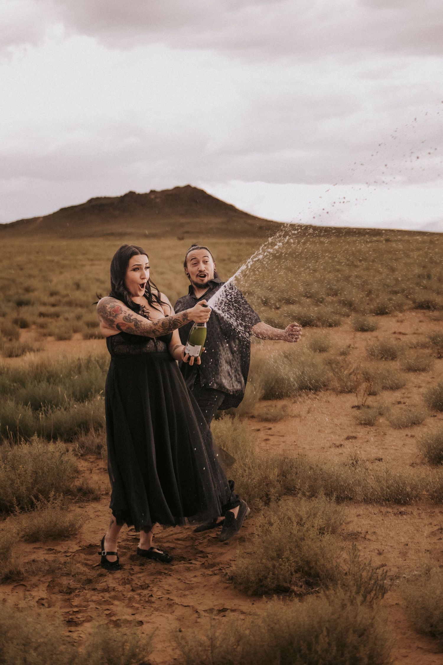 A couple in black dresses pops champagne together in a desert landscape with mountains in the background. New Mexico engagement photographer