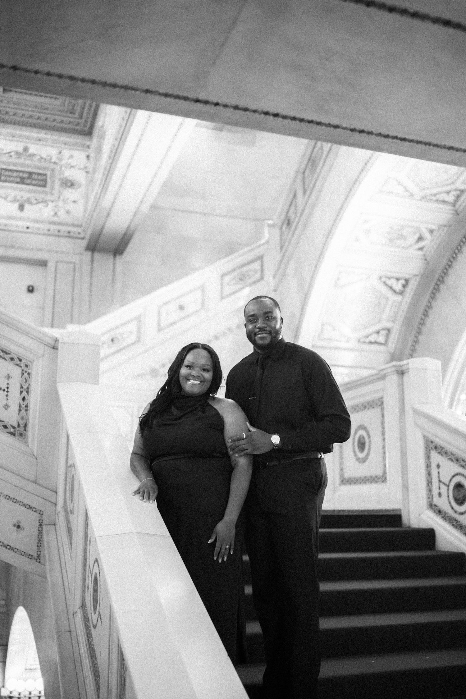 Couple in matching black outfits pose on elegant marble staircase with ornate architectural details in background.