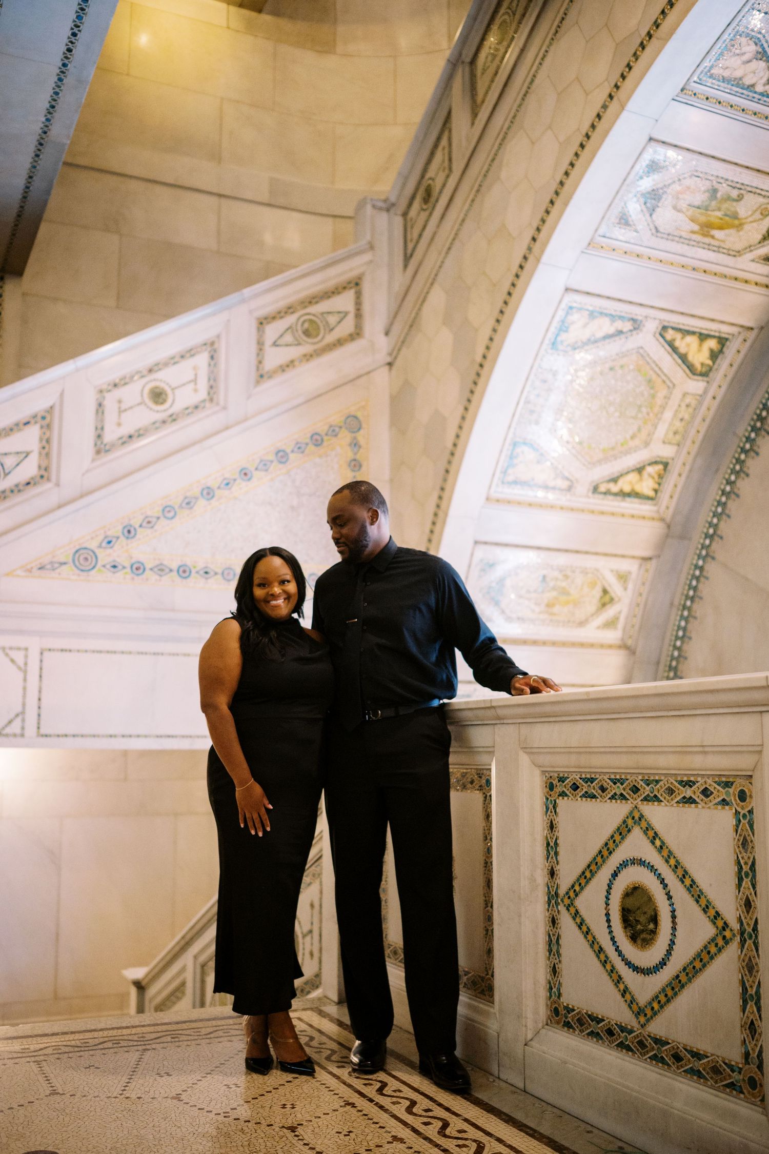 A couple stands lovingly together on an ornate marble staircase with decorative panels and architectural details.