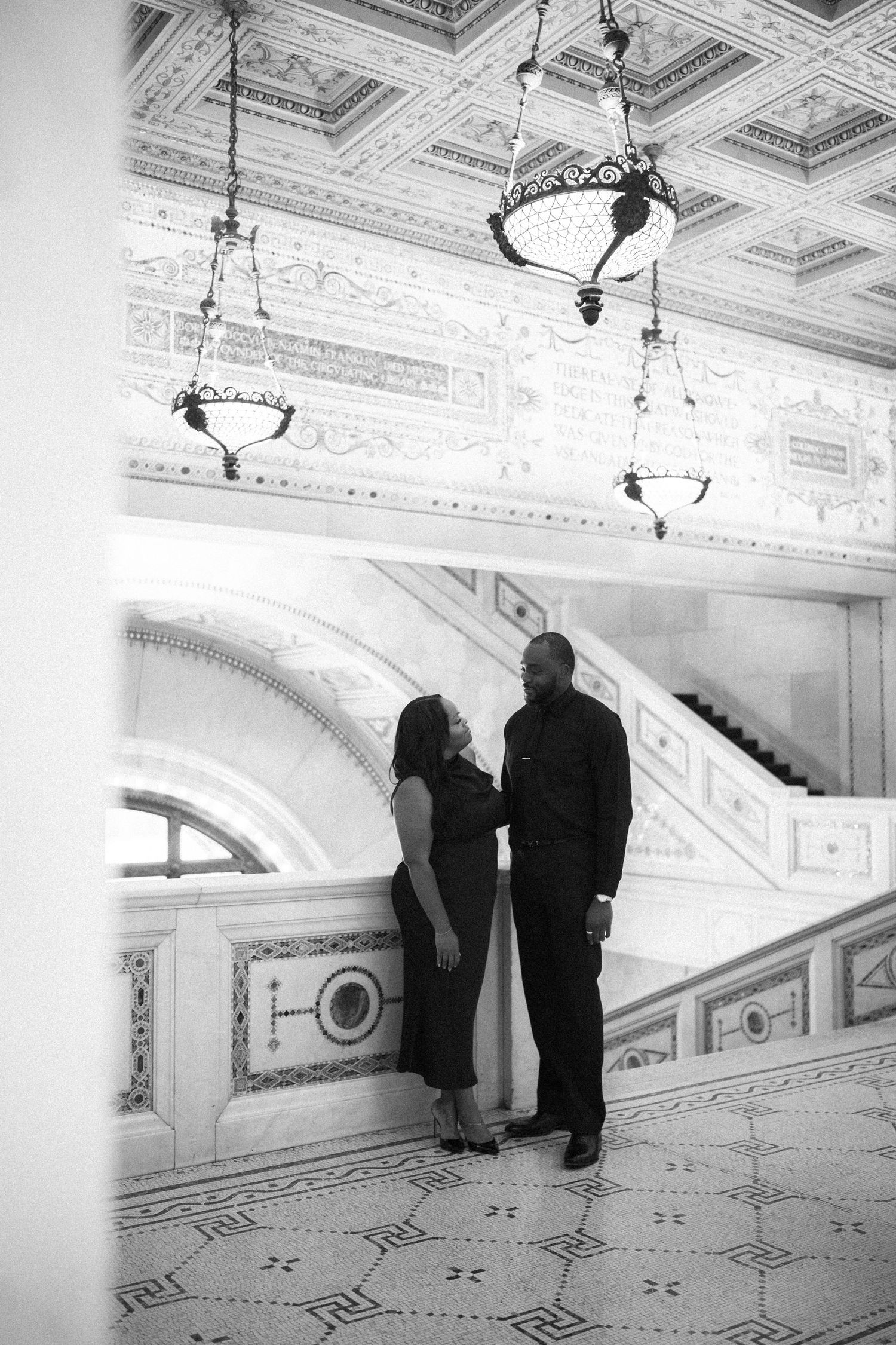 Romantic black and white portrait of couple beneath vintage chandeliers in historic building interior.