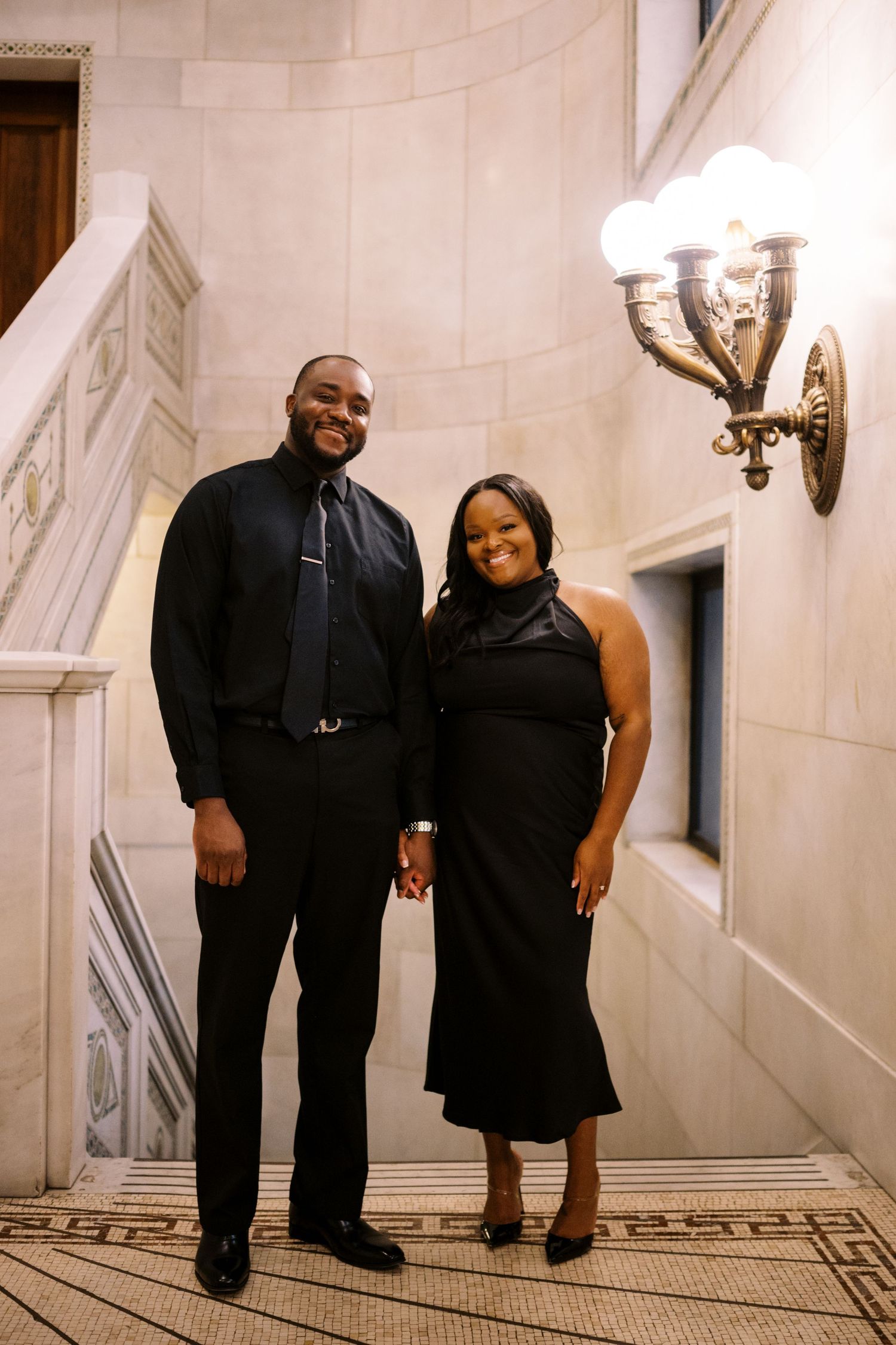Couple dressed in formal black attire pose together on marble staircase near decorative wall sconce.