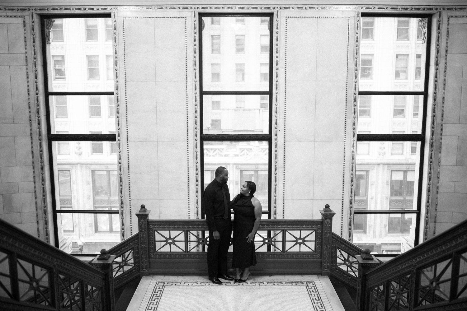 Two silhouettes stand in conversation by ornate window panels in a monochrome architectural interior.