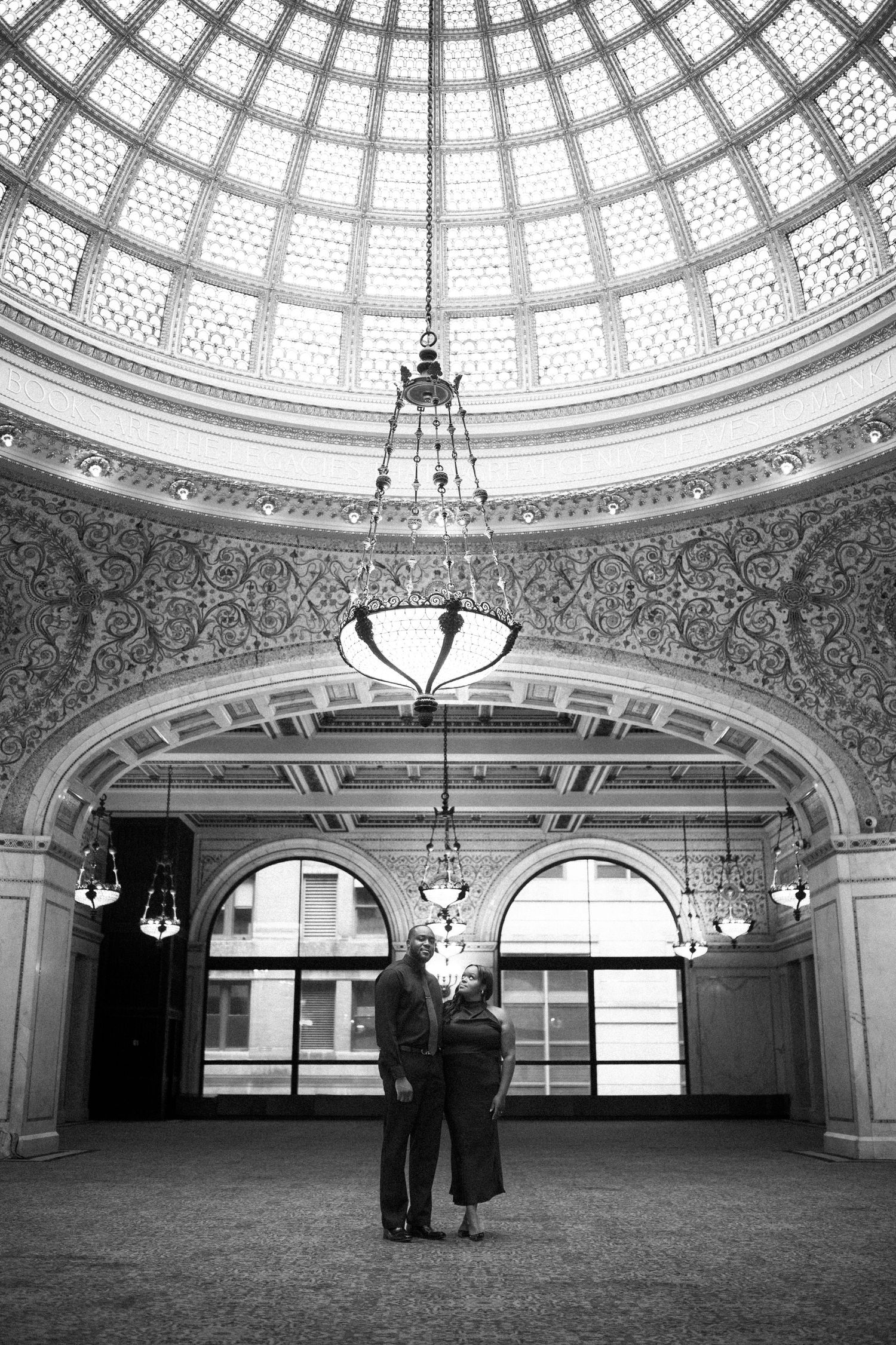 A couple stands beneath an ornate domed ceiling with intricate architectural details and hanging chandeliers.