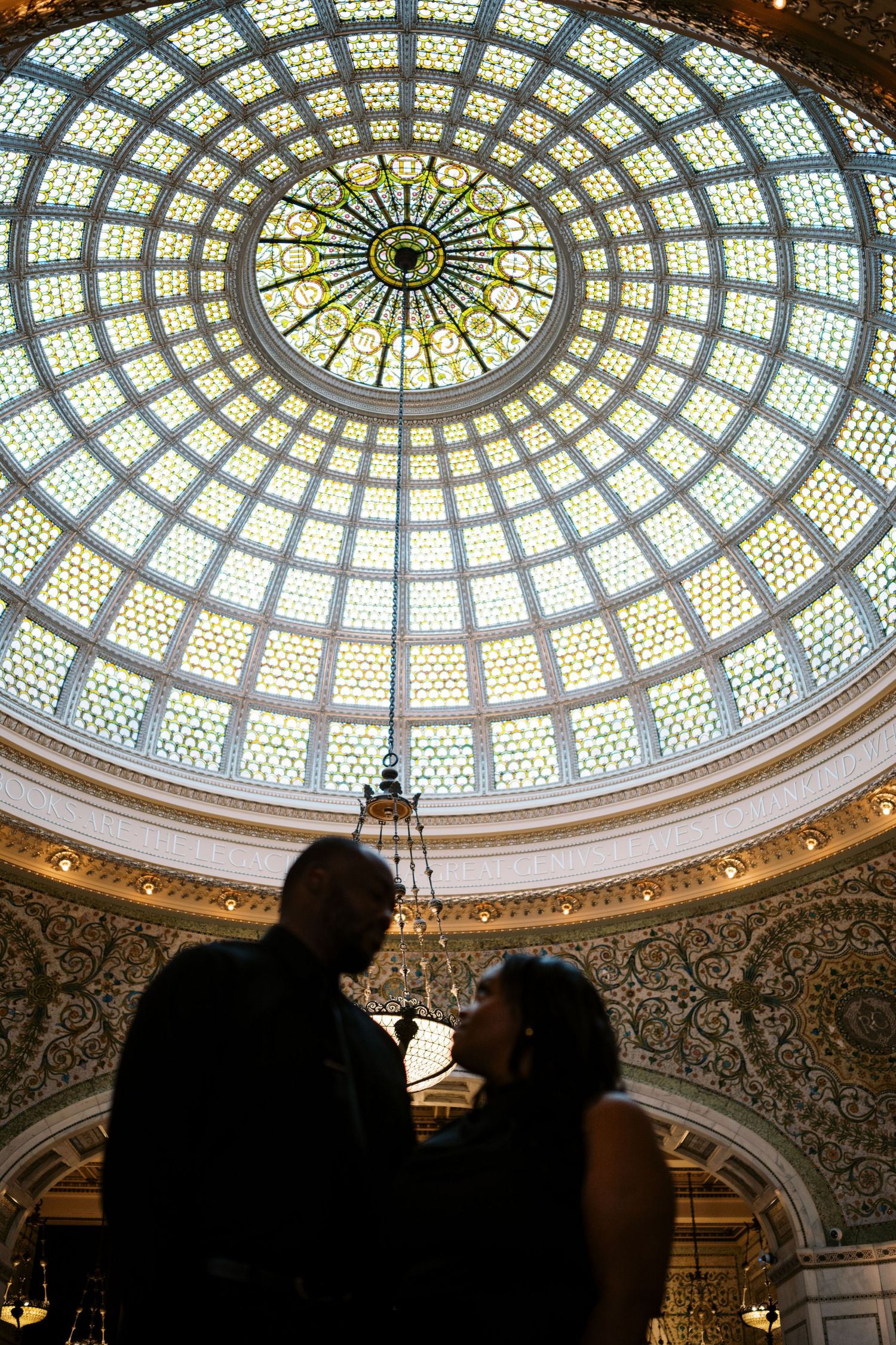 A silhouetted couple embraces beneath an ornate stained glass dome ceiling with intricate geometric patterns.