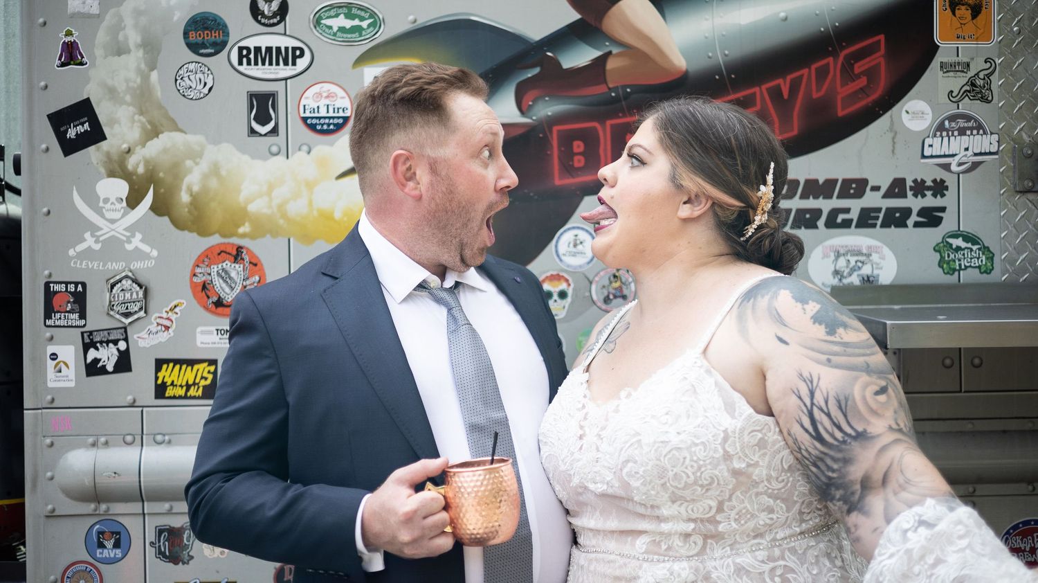 A bride and groom share a joyful moment while holding drinks in front of a wall covered in stickers and street art.