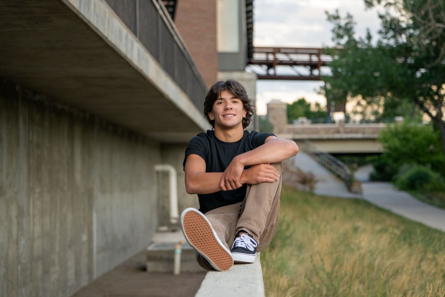 A young person sits casually on a low wall outdoors wearing a black t-shirt and sneakers.