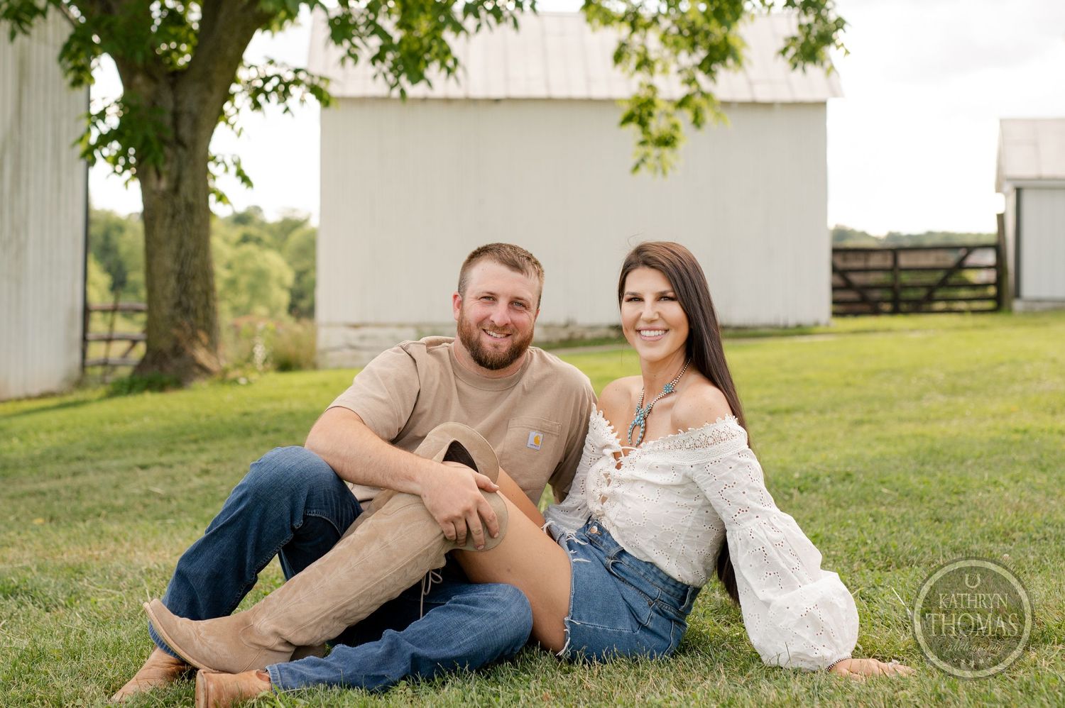 Couple sits together on grassy hill with white barn in background during outdoor photoshoot.