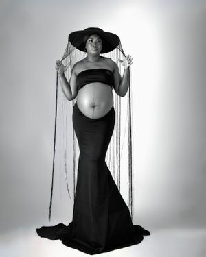 a black and white photograph of pregnant lady in a black dress and a hat