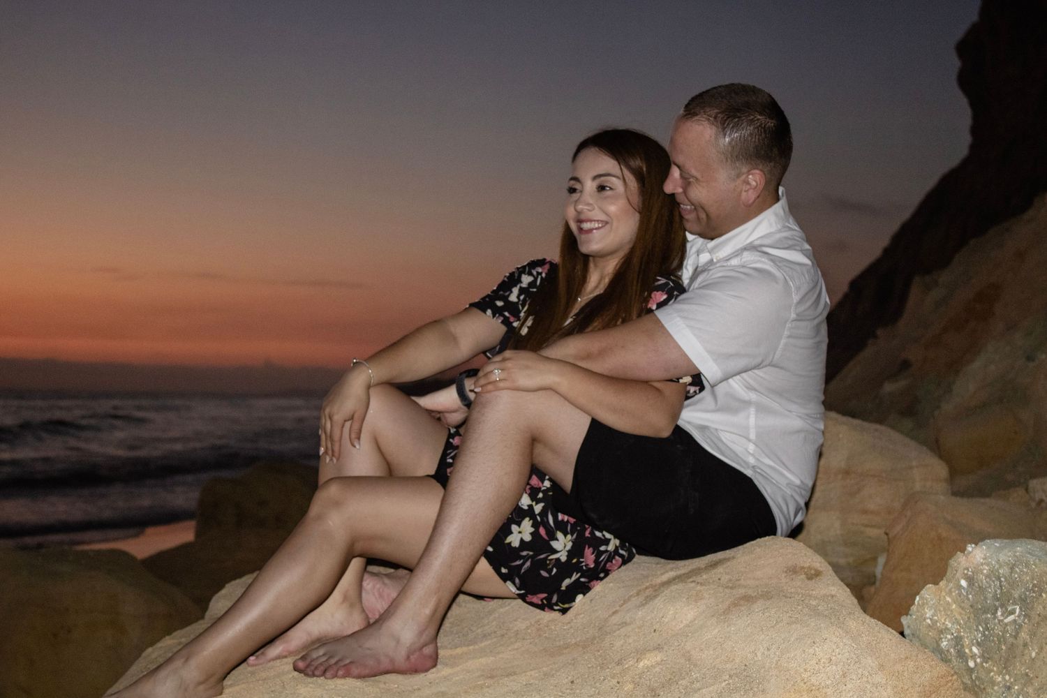 Couple sitting together on coastal rocks during a dramatic sunset.