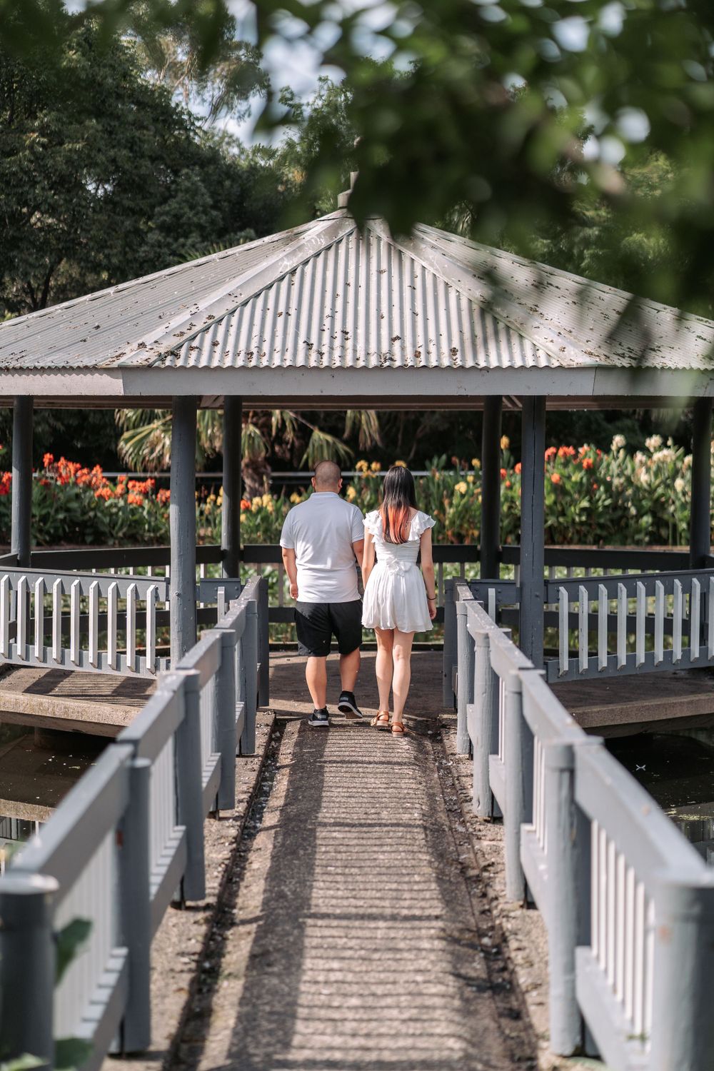 Couple walking towards gazebo on wooden bridge pathway with white railings.