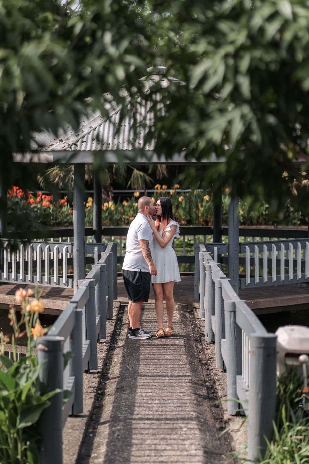 Couple sharing intimate moment on wooden bridge pathway surrounded by flowers.