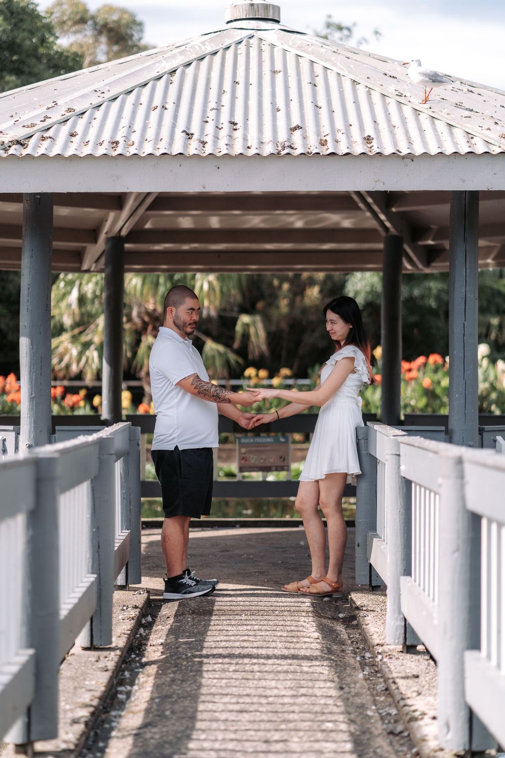 A couple stands facing each other on a covered wooden bridge in a scenic garden.