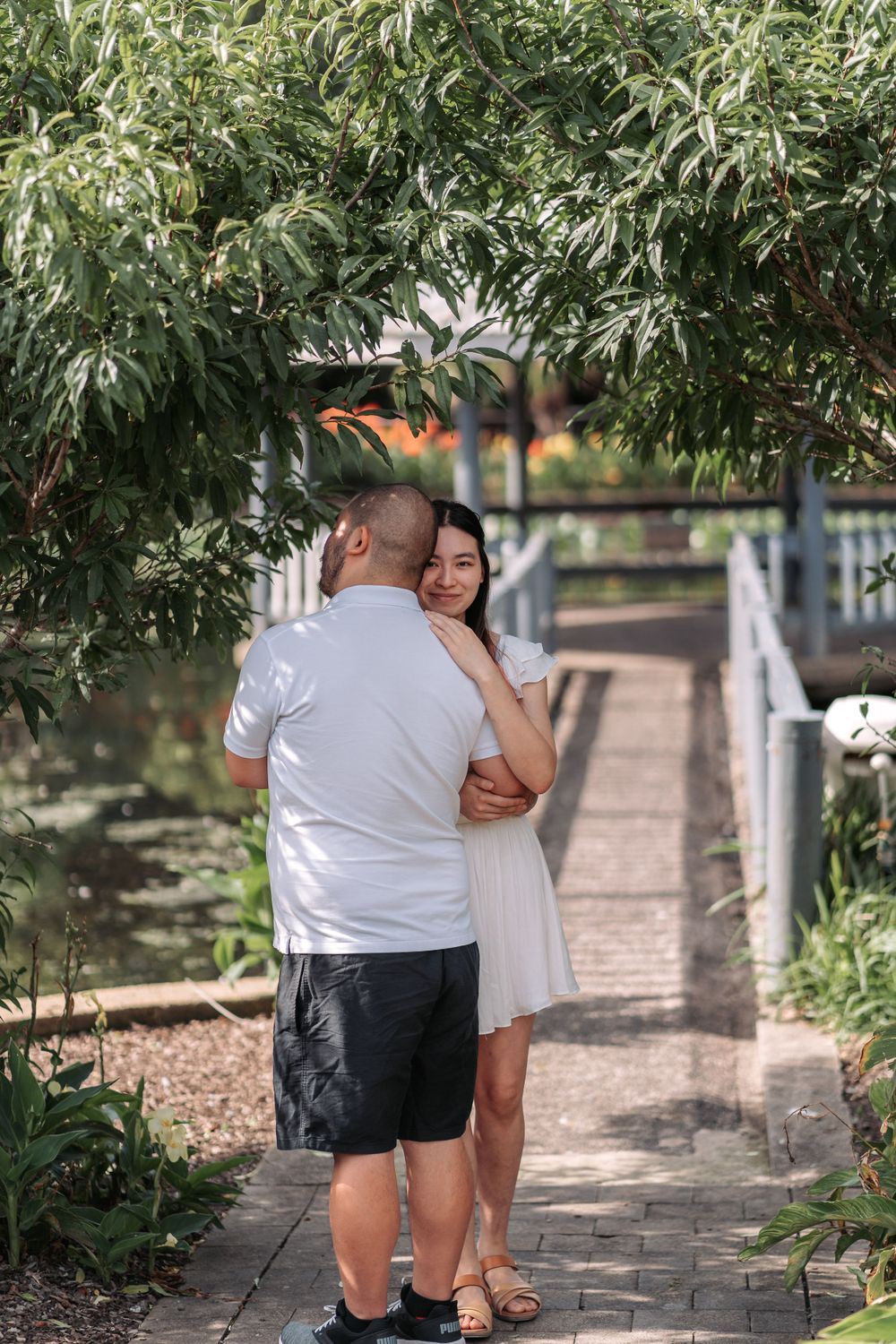A couple embraces on a garden path surrounded by overhanging trees and landscaping.