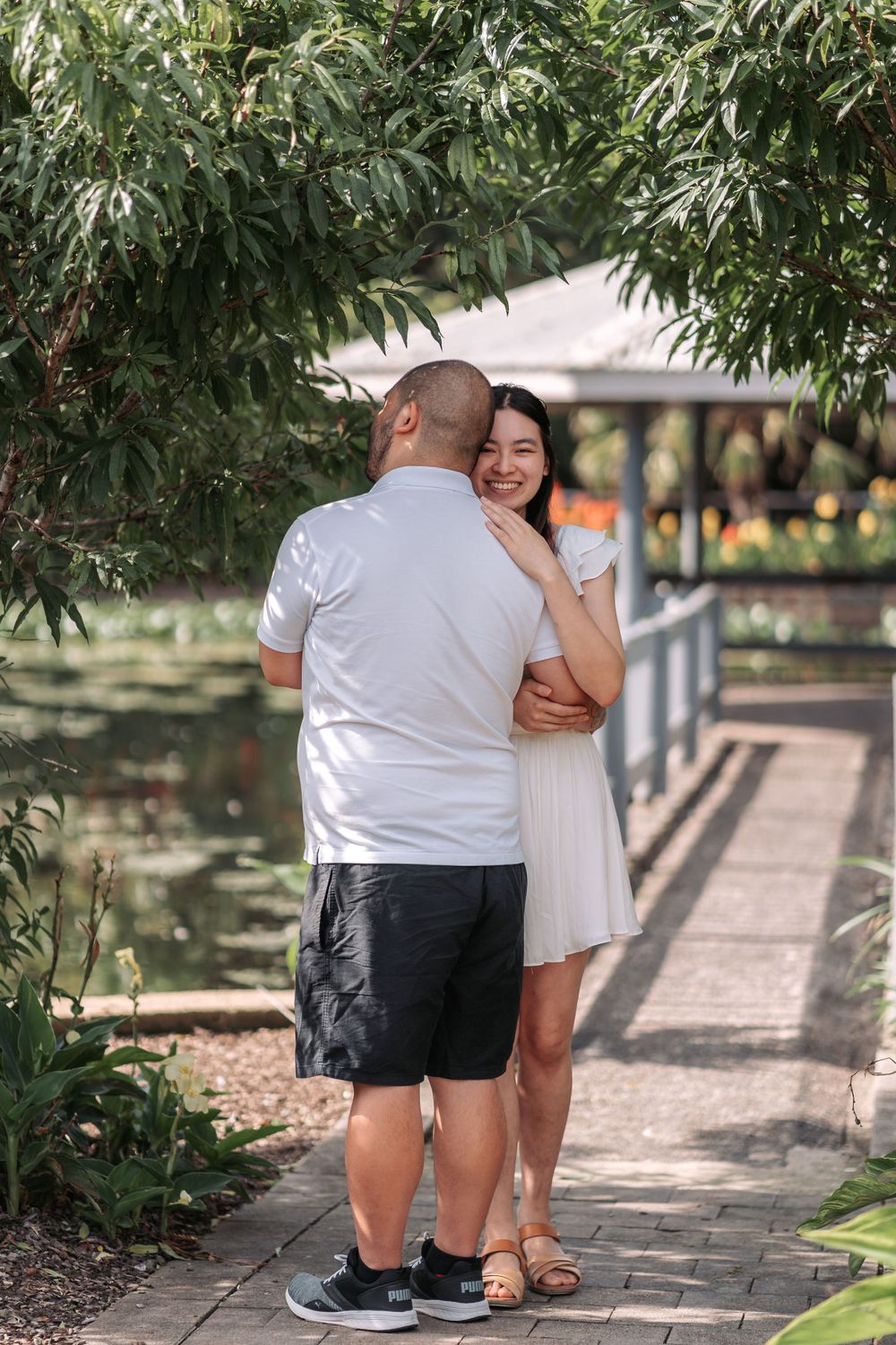A couple shares an intimate moment on a brick pathway under leafy green trees.