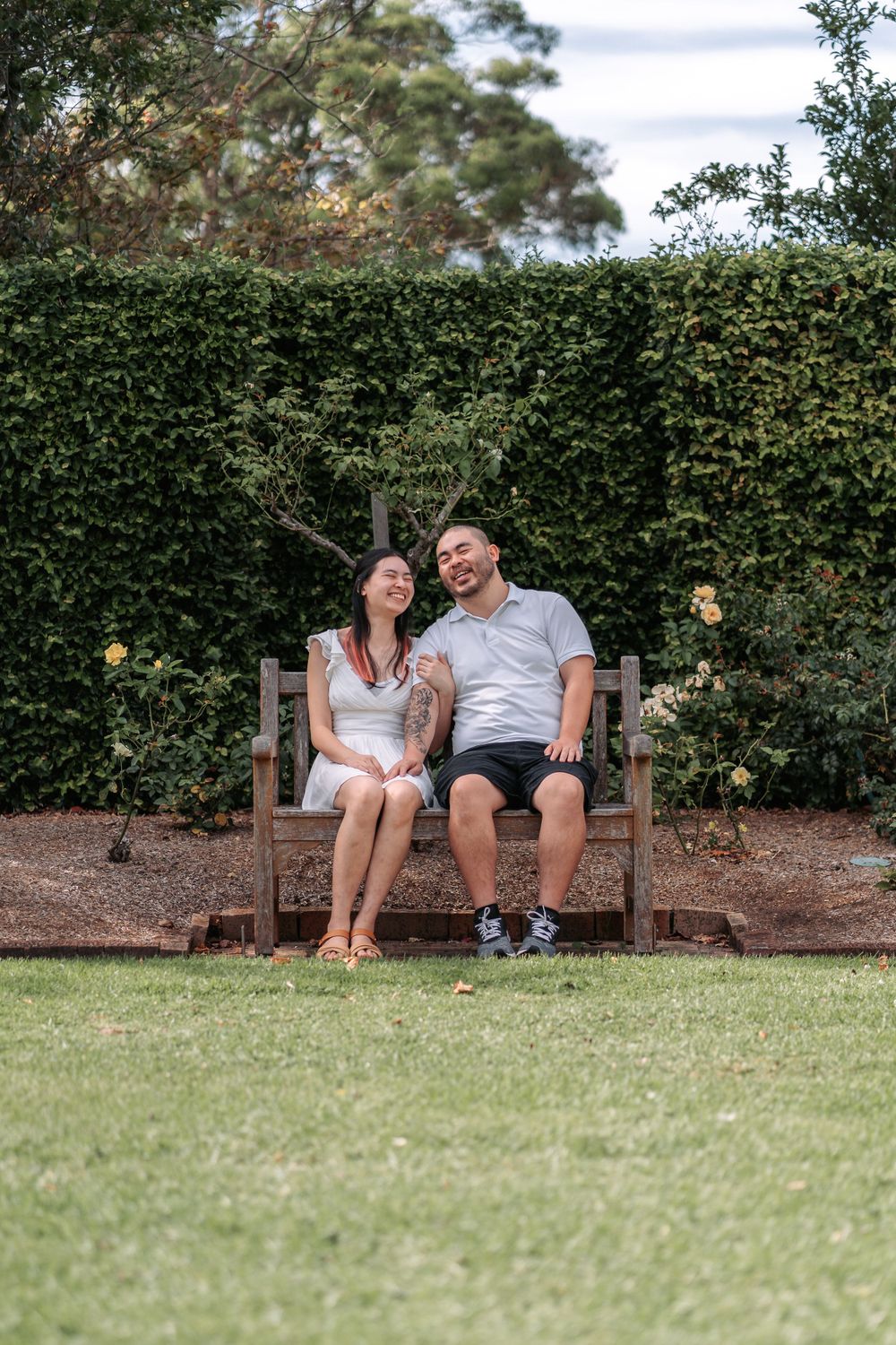 A couple sitting together on a wooden bench against a manicured hedge in a garden setting.