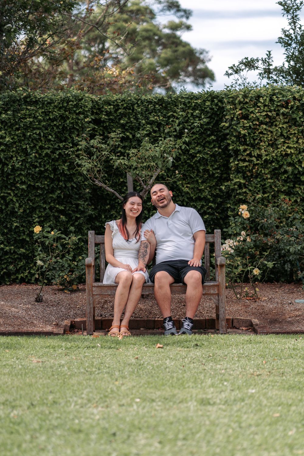 A couple laughing together while seated on a wooden garden bench surrounded by hedges.