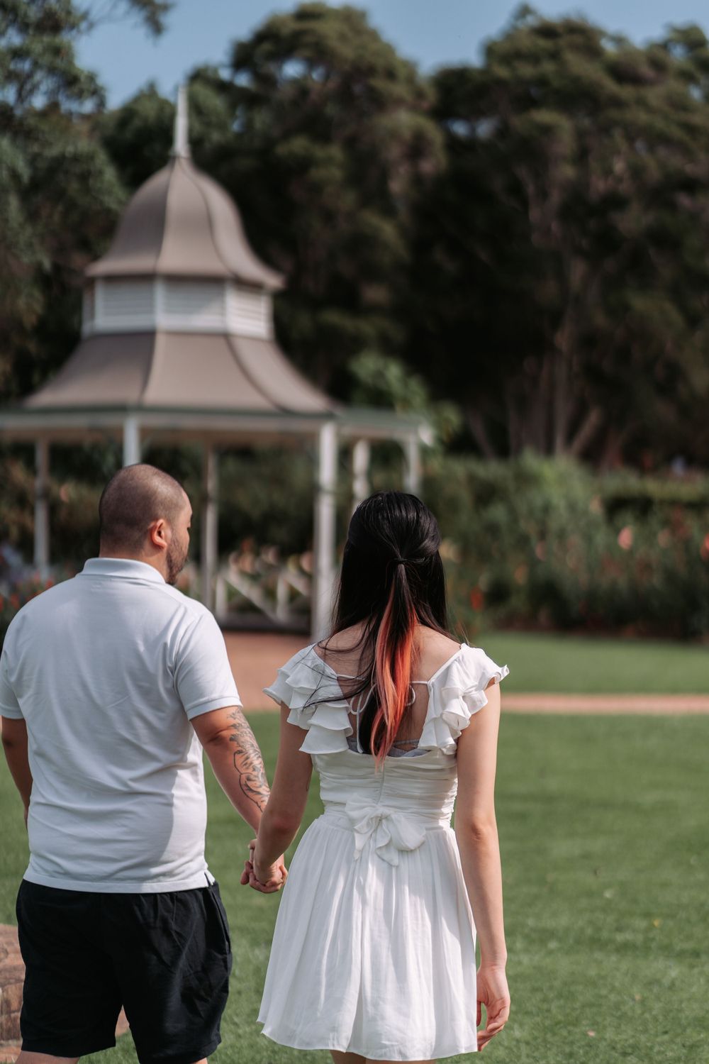A couple walking hand in hand towards a gazebo in a lush garden setting.
