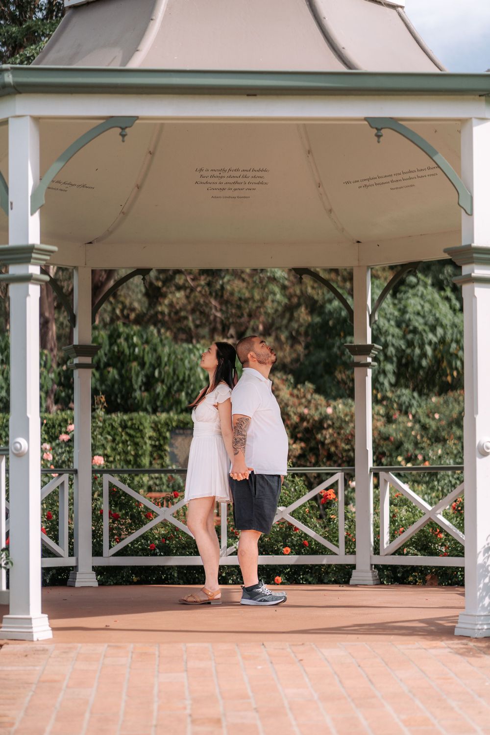 Elegant white gazebo with ornate railings surrounded by blooming garden flowers and trees.