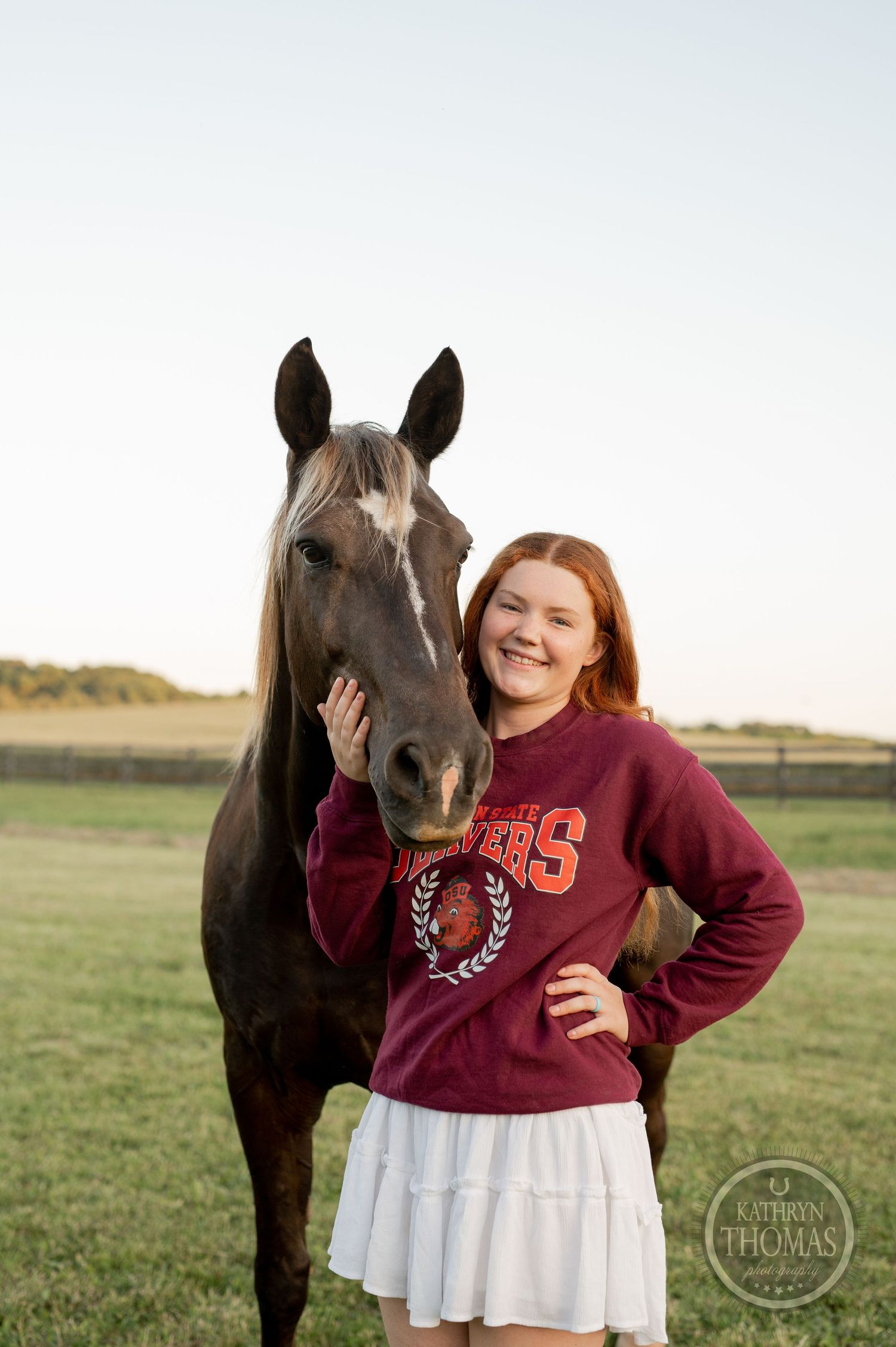 A dark horse and person in a burgundy sweatshirt share a tender moment in a grassy field at sunset.