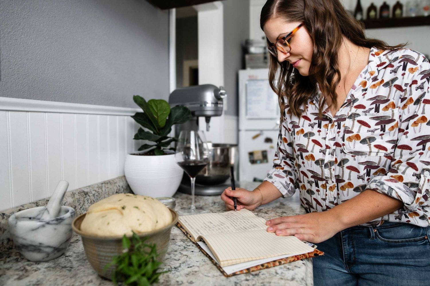 A person in a patterned shirt writes in a notebook while standing at a kitchen counter.