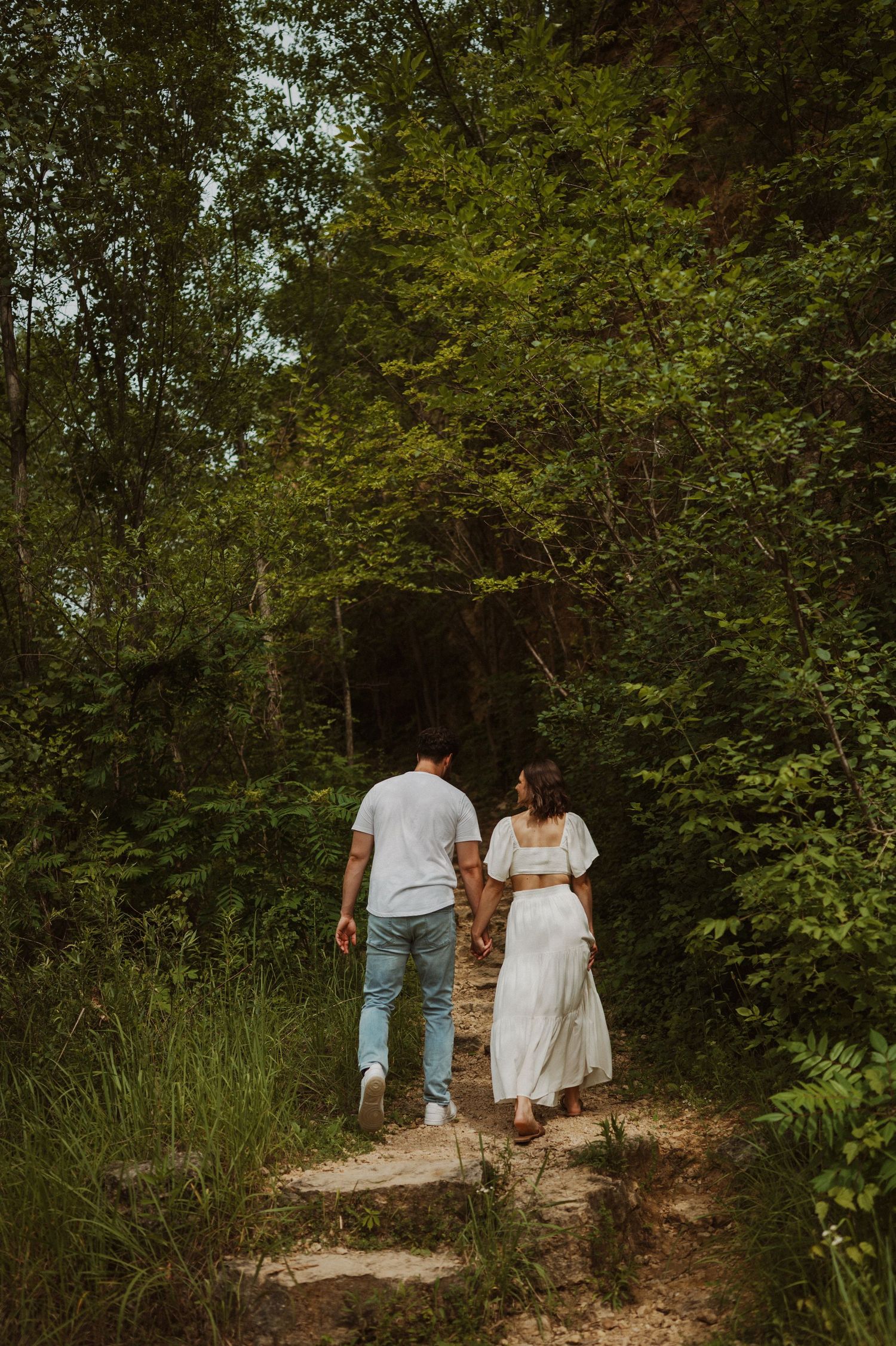 A couple walks hand in hand up stone steps on a wooded trail.