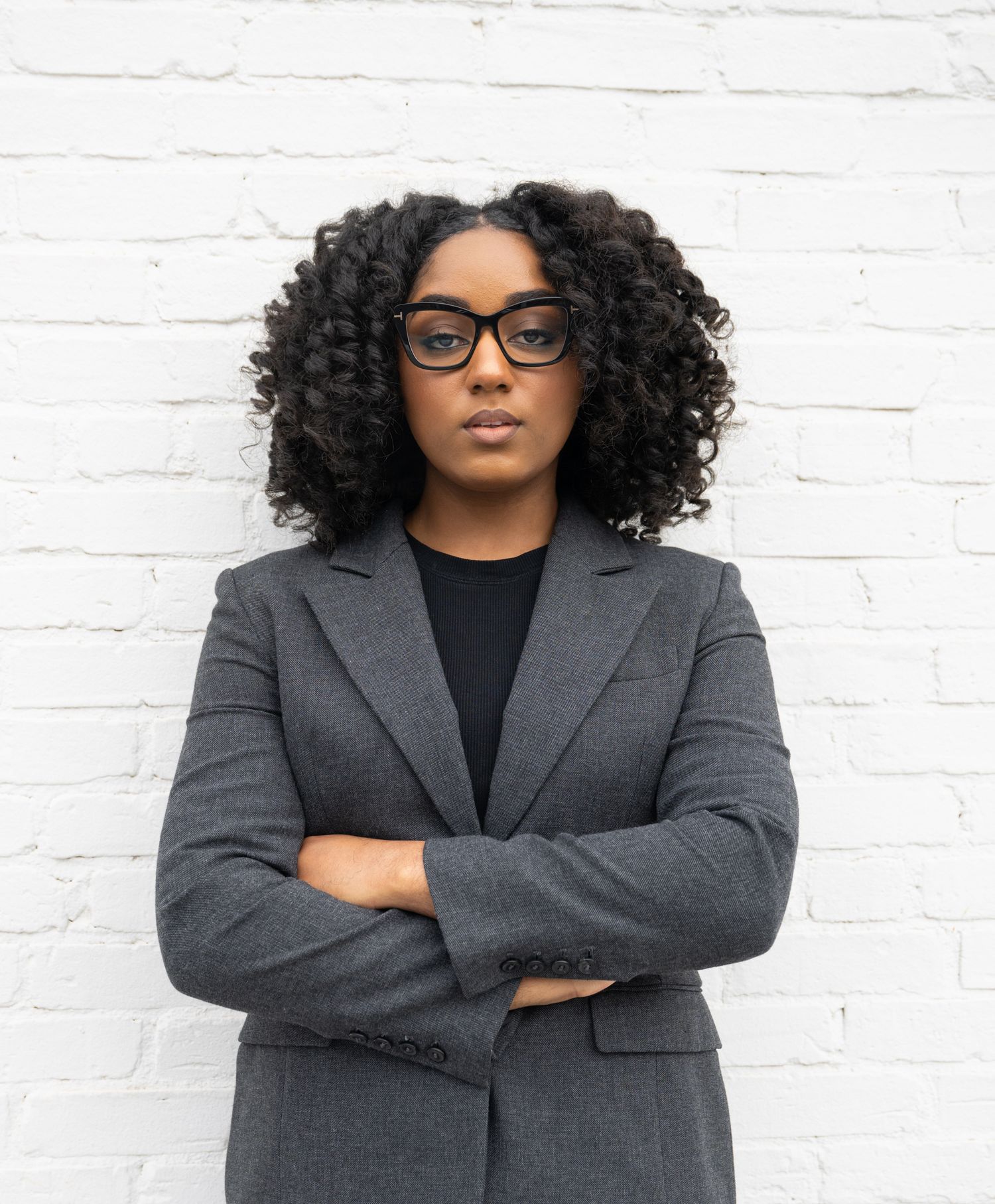 Reyna Waters wearing a gray blazer and black top stands confidently with arms crossed against a white brick wall.