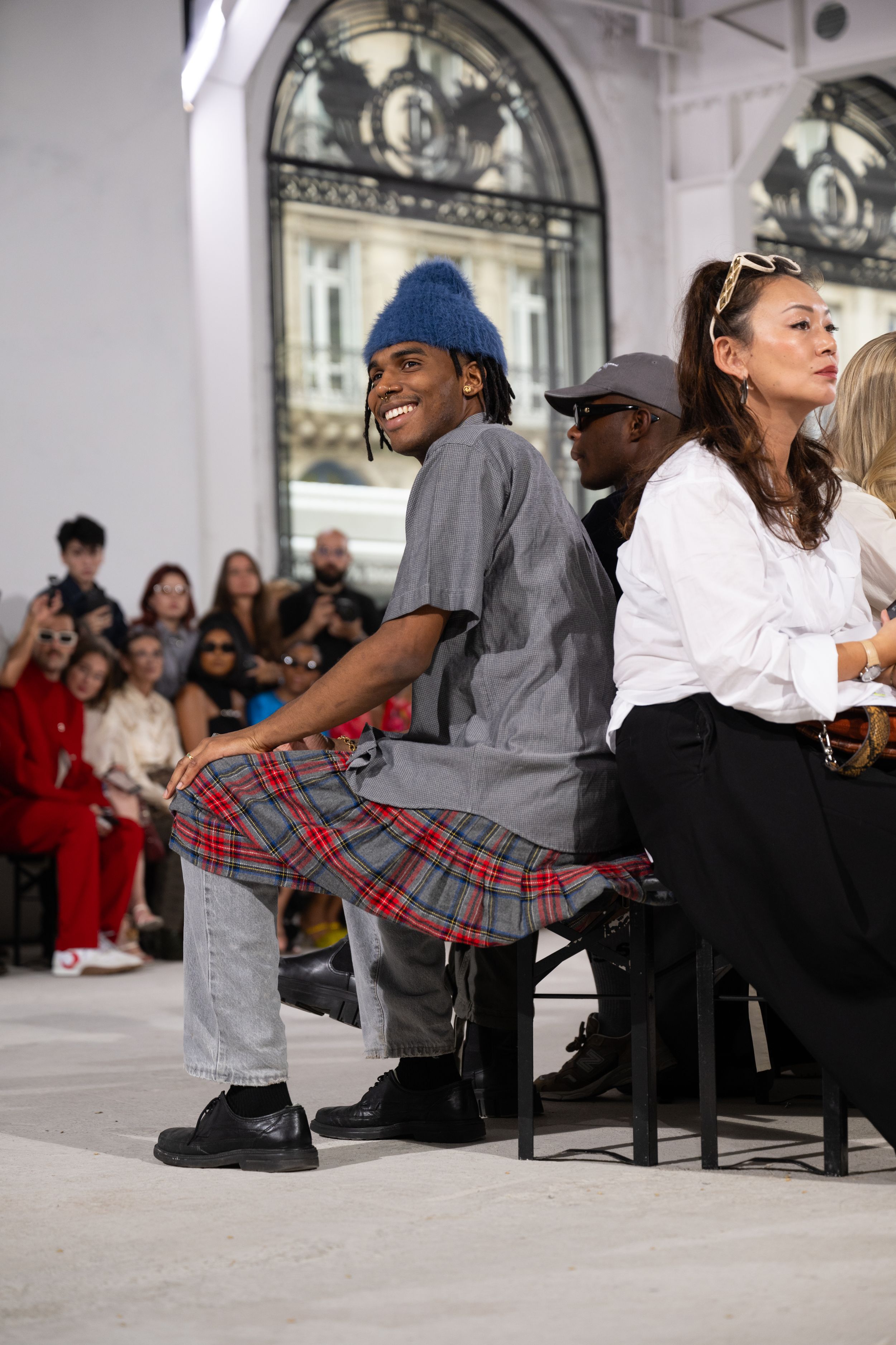 Fashion show attendee in casual plaid skirt and beanie seated among audience members.