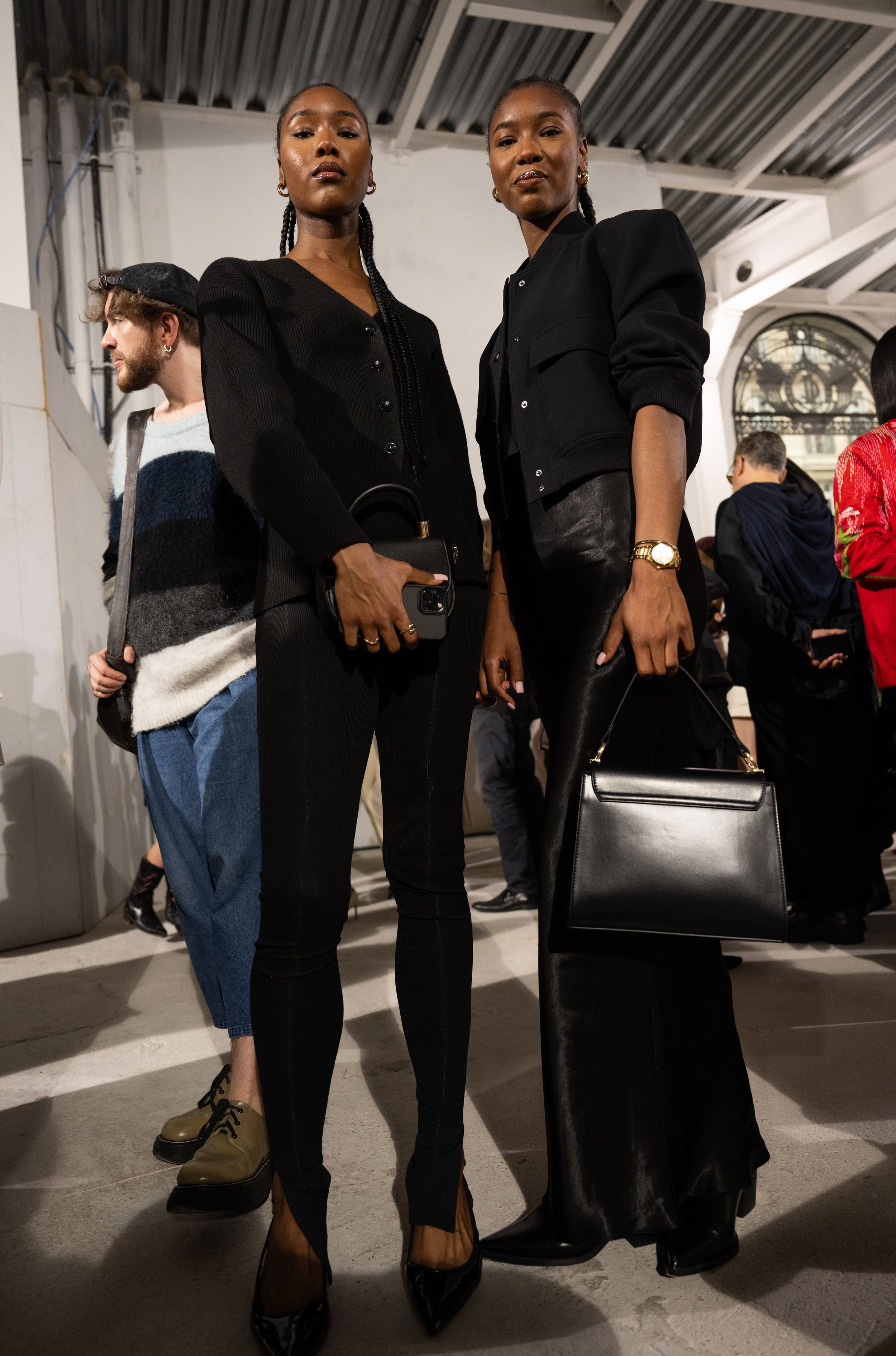 Two models in coordinated all-black outfits with structured handbags at fashion event.