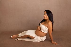 A person in a white dress poses gracefully on the floor during a maternity photo session with a brown backdrop. Sarah Jane Photography LLC
