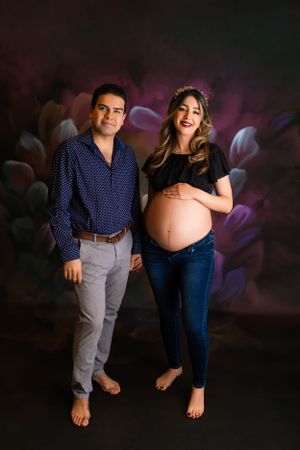A couple stands barefoot against a dark floral backdrop during a maternity photoshoot. Maternity & Newborn Photographer in NJ