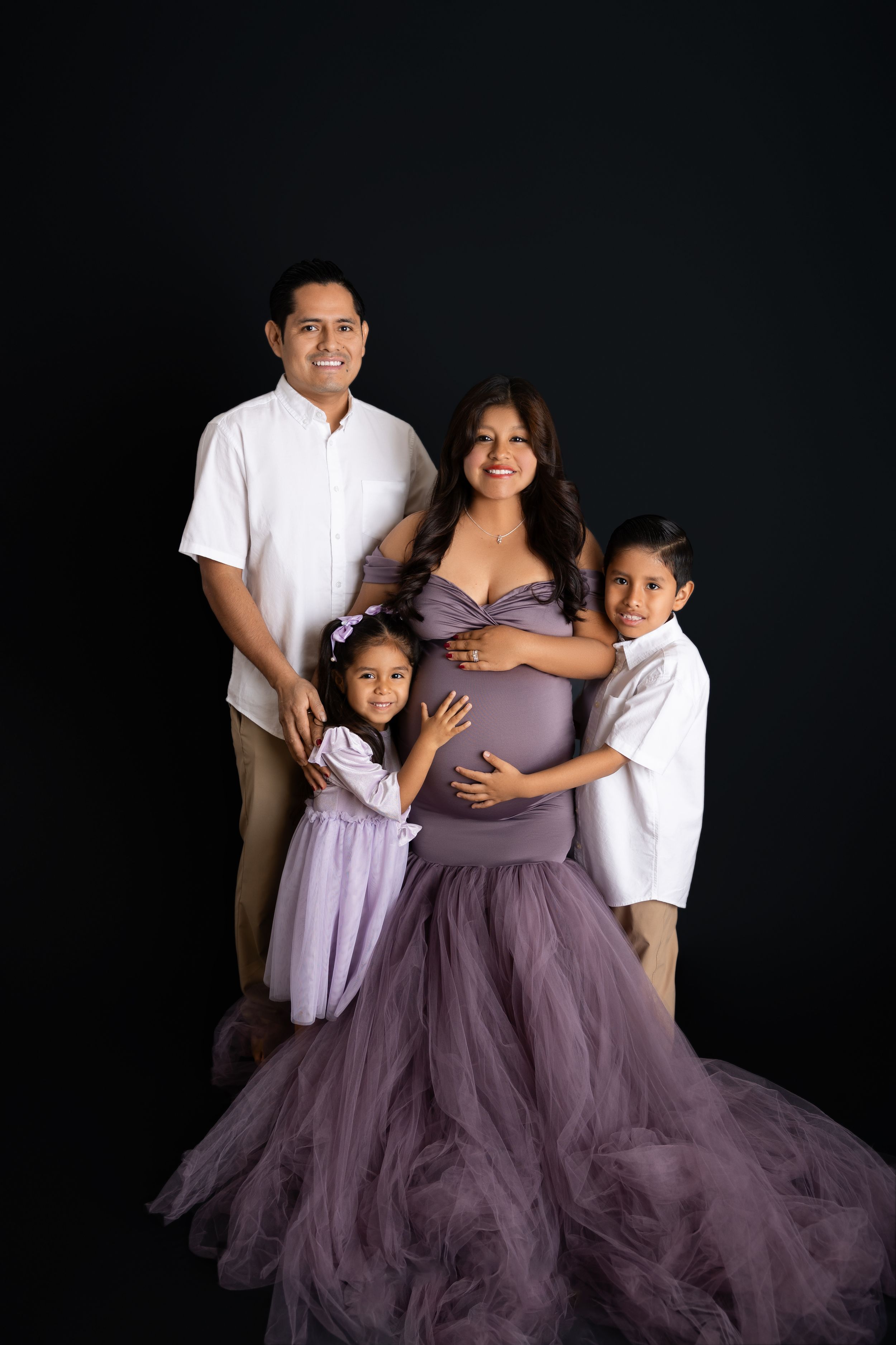 A family wearing matching purple tulle outfits poses for elegant portrait photos against a dark background. NJ MATERNITY PHOTOGRAPHY