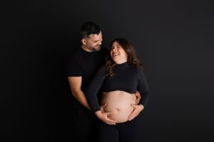 A couple in black attire stands against dark backdrop during maternity photoshoot, sweetly embracing the baby bump. Newton, NJ