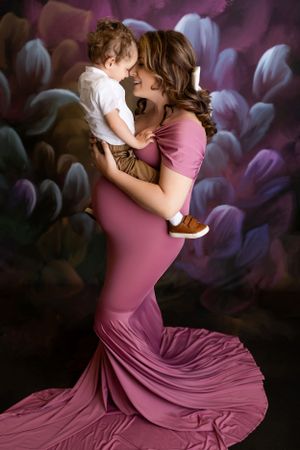 A touching portrait of an embrace against a dark floral backdrop with figure in flowing mauve gown holding small child. - NJ Photographer