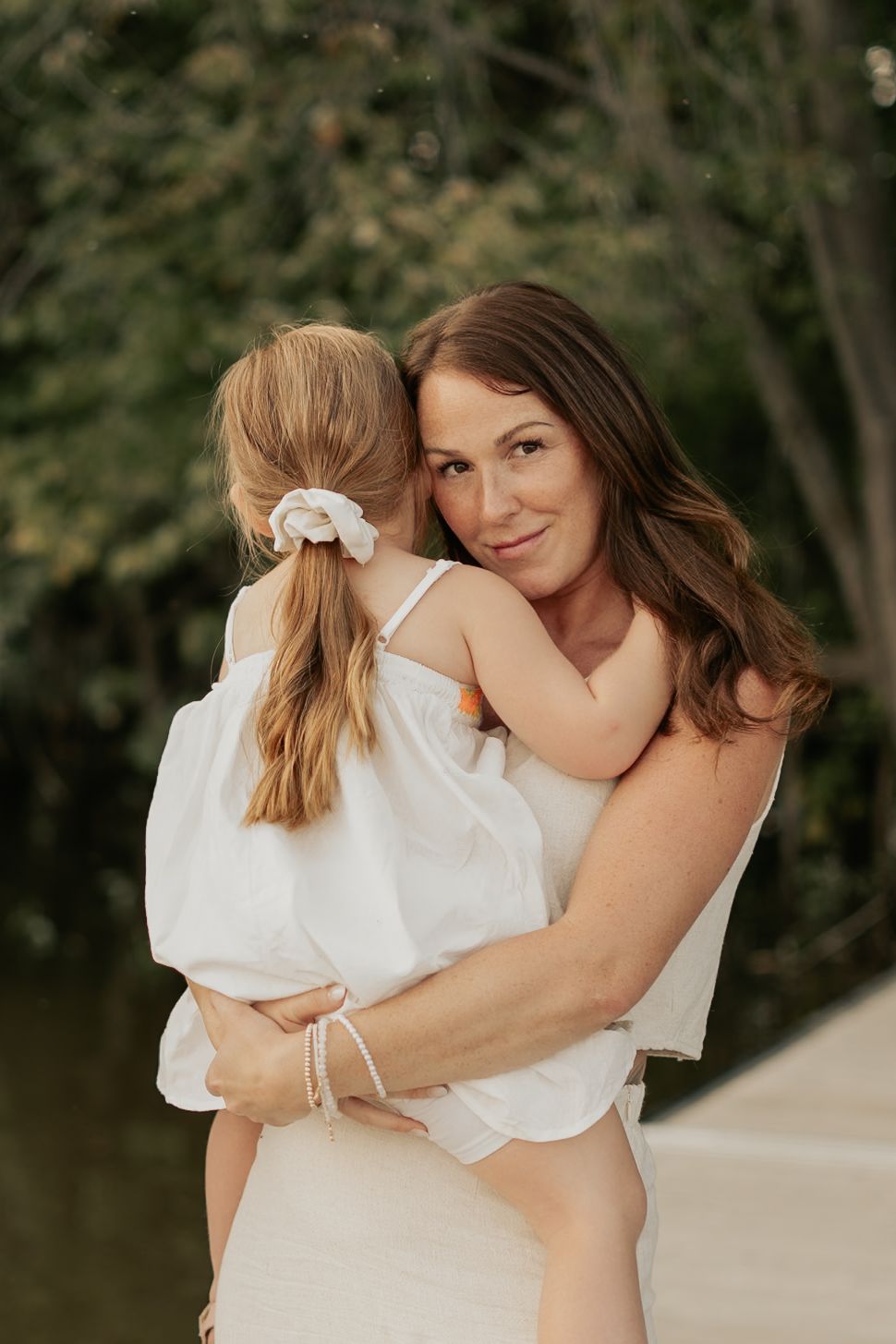 A tender moment outdoors as mother and daughter in white outfits embrace while surrounded by greenery.