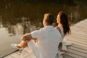 A couple in white outfits sits together on a wooden dock during golden sunset hour by a calm lake.