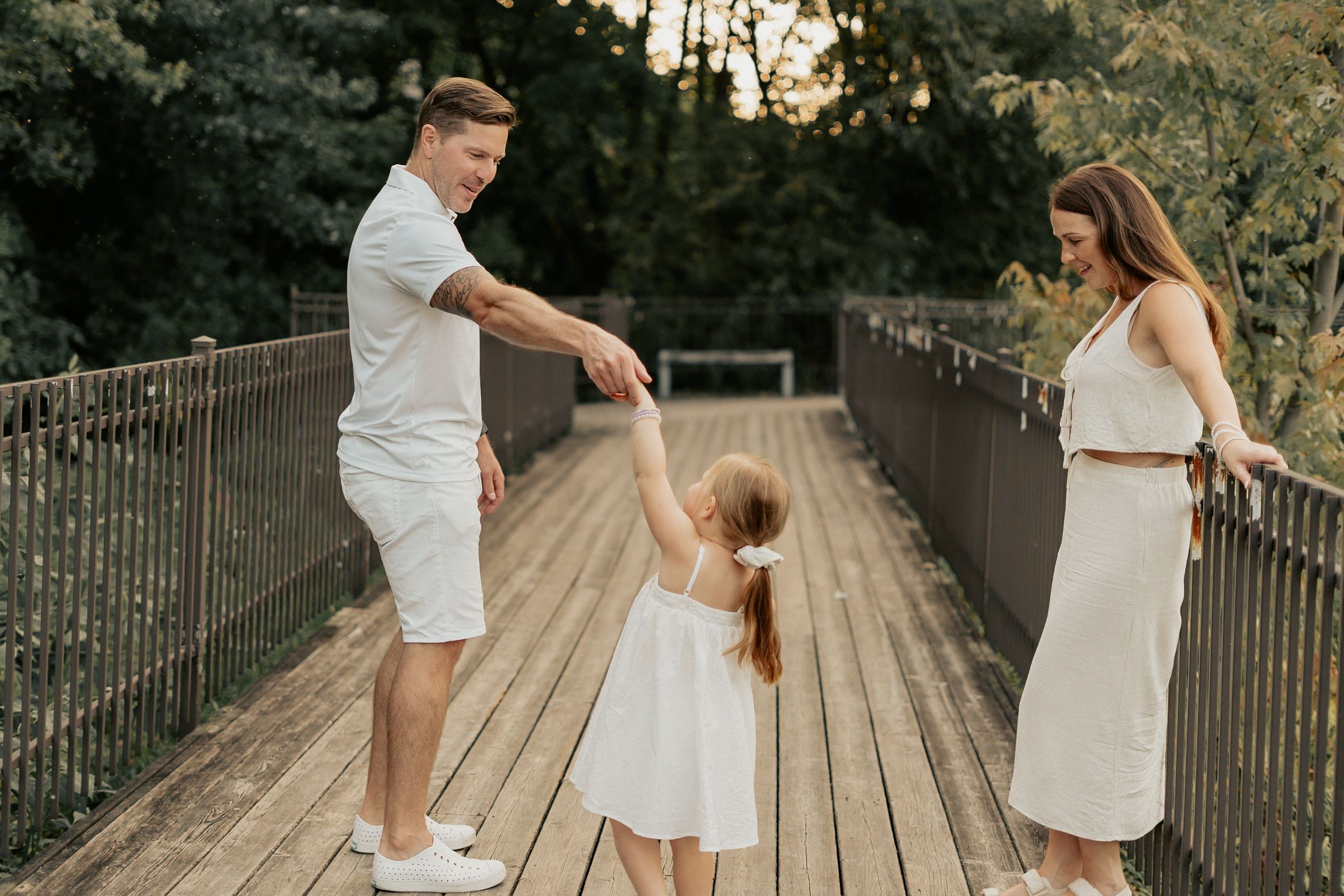 A family in white outfits holds hands and dances together on a wooden bridge during golden hour.