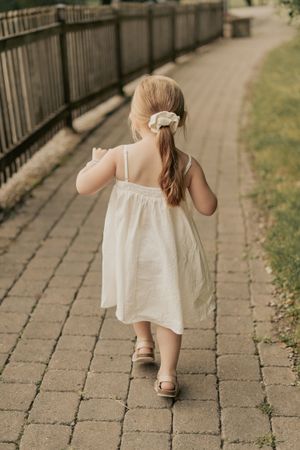 A small child in a white summer dress walks along a brick pathway bordered by a wooden fence on a sunny day.