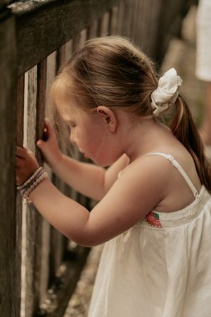 A young child in a white dress with a bow in their hair peeks through an old wooden fence in soft lighting.
