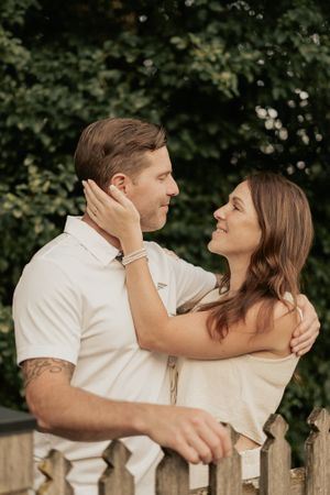 Two people in light colored outfits embrace tenderly while standing by a wooden fence.
