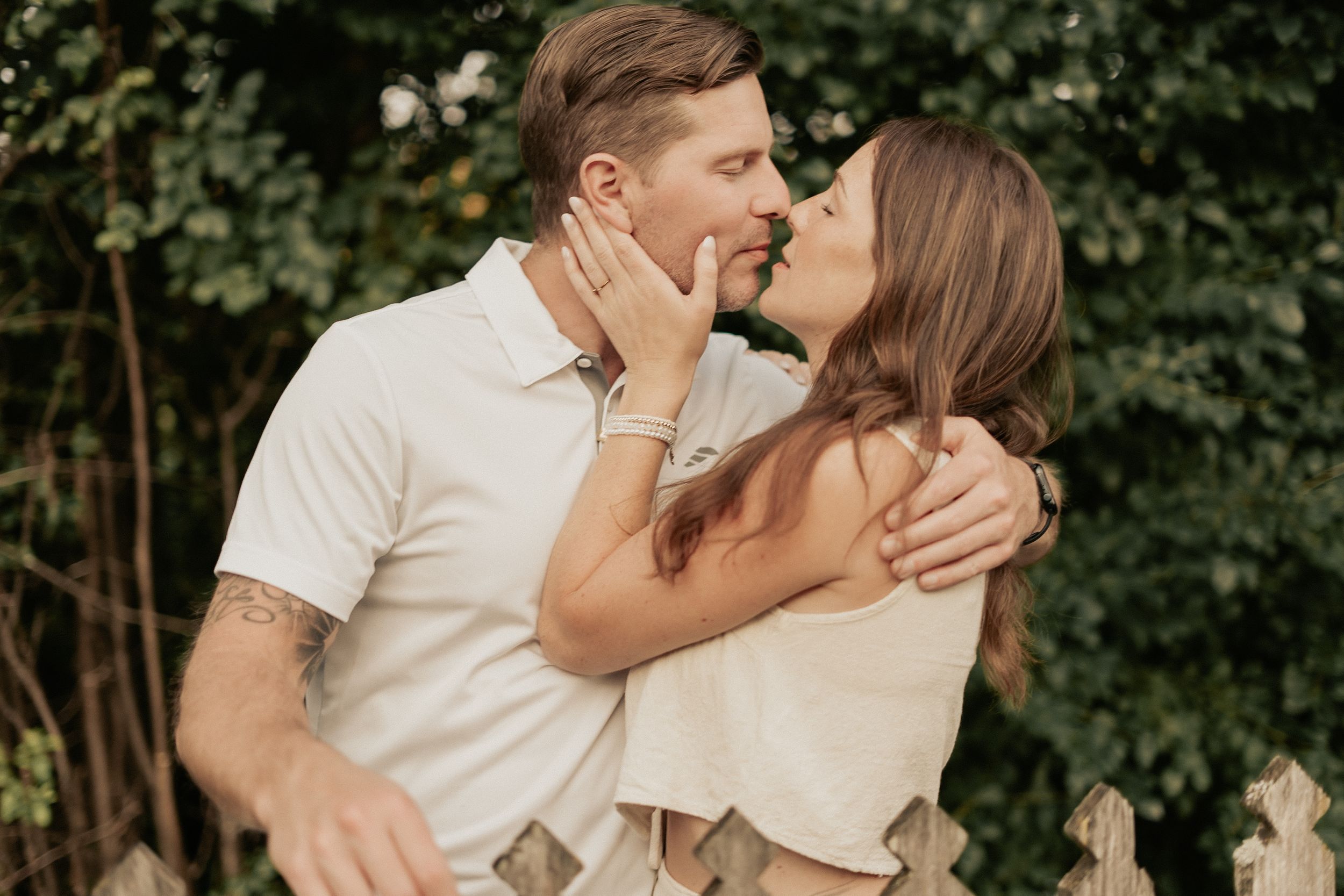 A romantic couple sharing a tender kiss outdoors with green foliage in the background.
