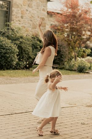 Two people in light colored dresses dance joyfully together on a stone patio near a garden with autumn foliage.