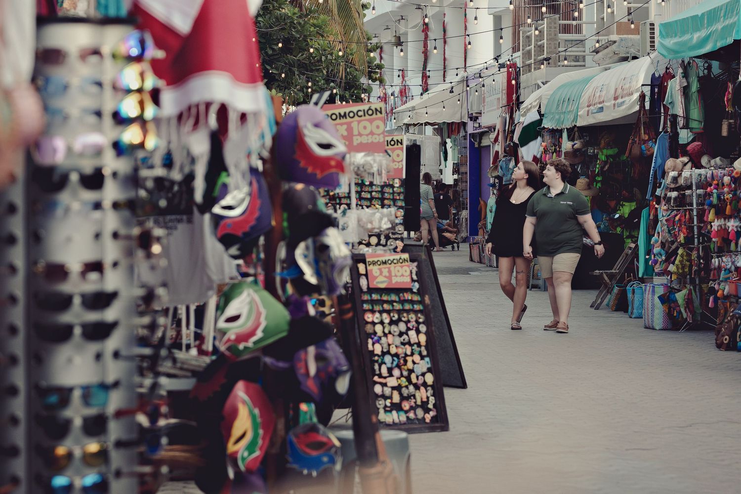 Outdoor market street with souvenir stands displaying colorful merchandise and sunglasses along a busy sidewalk.