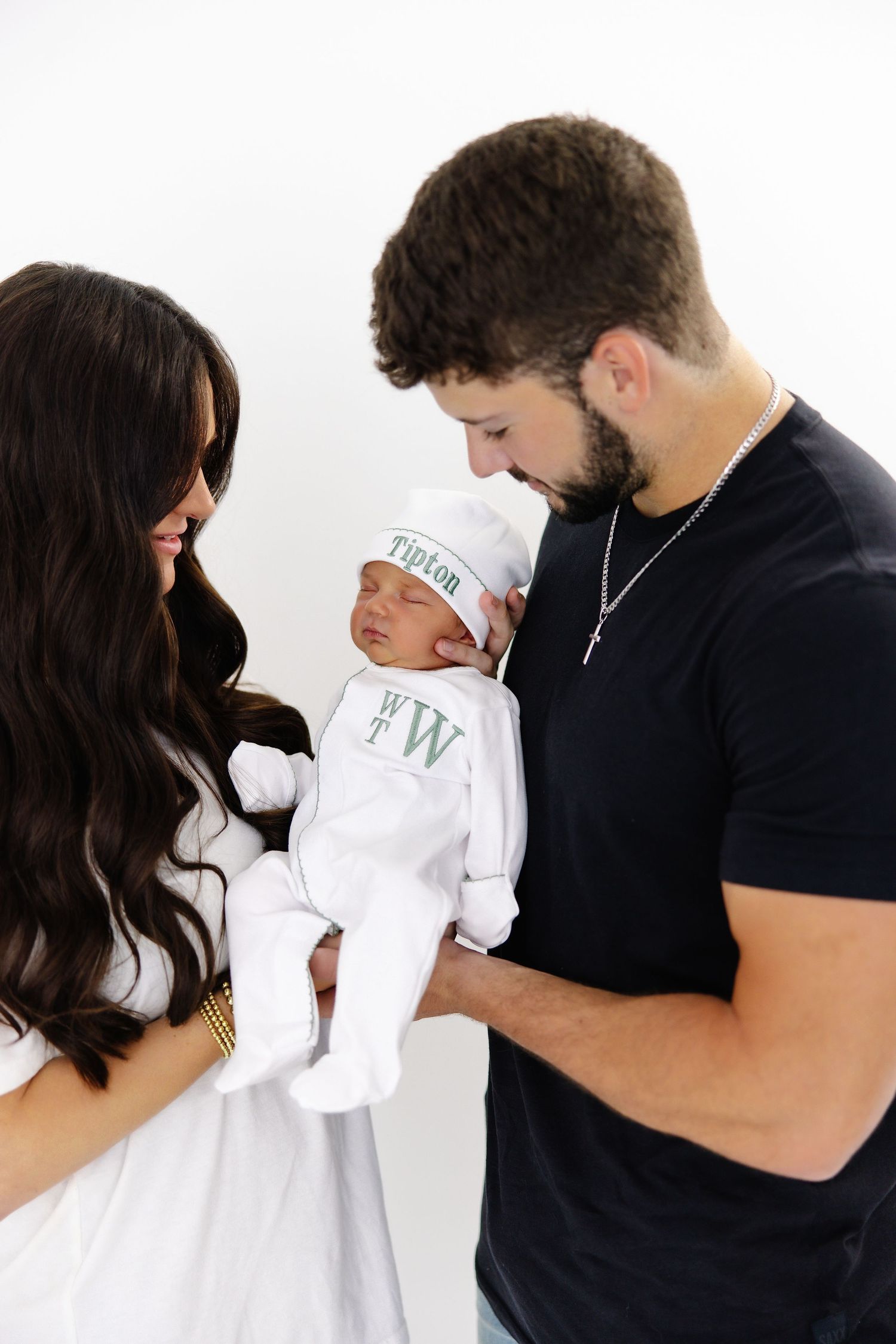 Parents hold their newborn wrapped in a white Tipton branded swaddle against a white background.