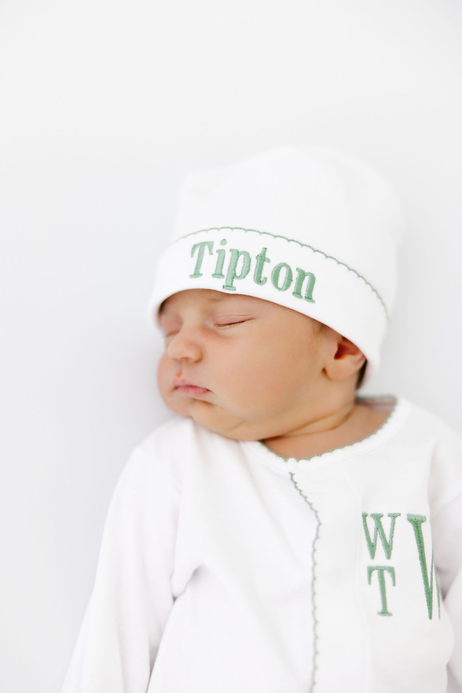 Sleeping newborn baby wearing a white hat with green Tipton lettering on a white background.