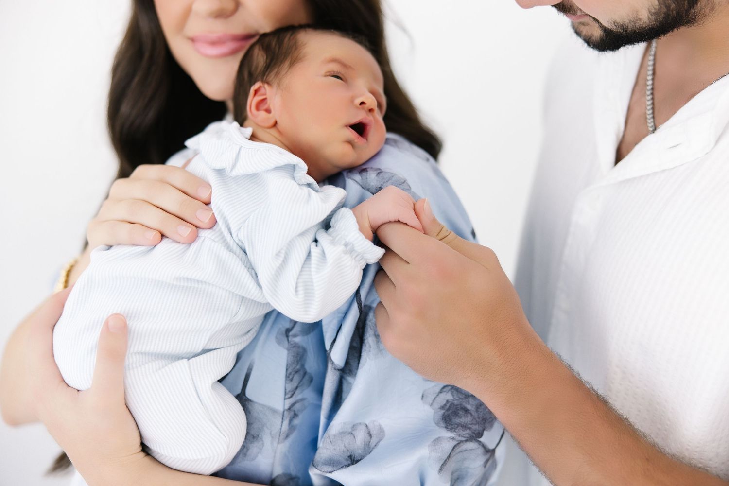 Family snuggles and admires their newborn baby wearing white clothing against neutral backdrop.