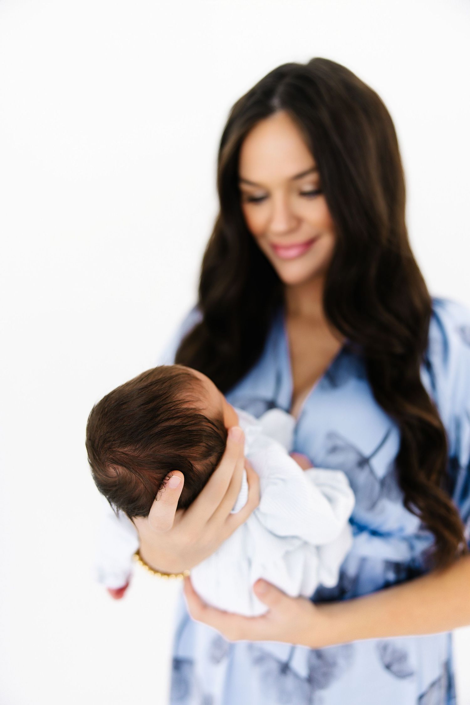 A woman in a blue floral dress cradles a sleeping newborn baby in a white outfit.