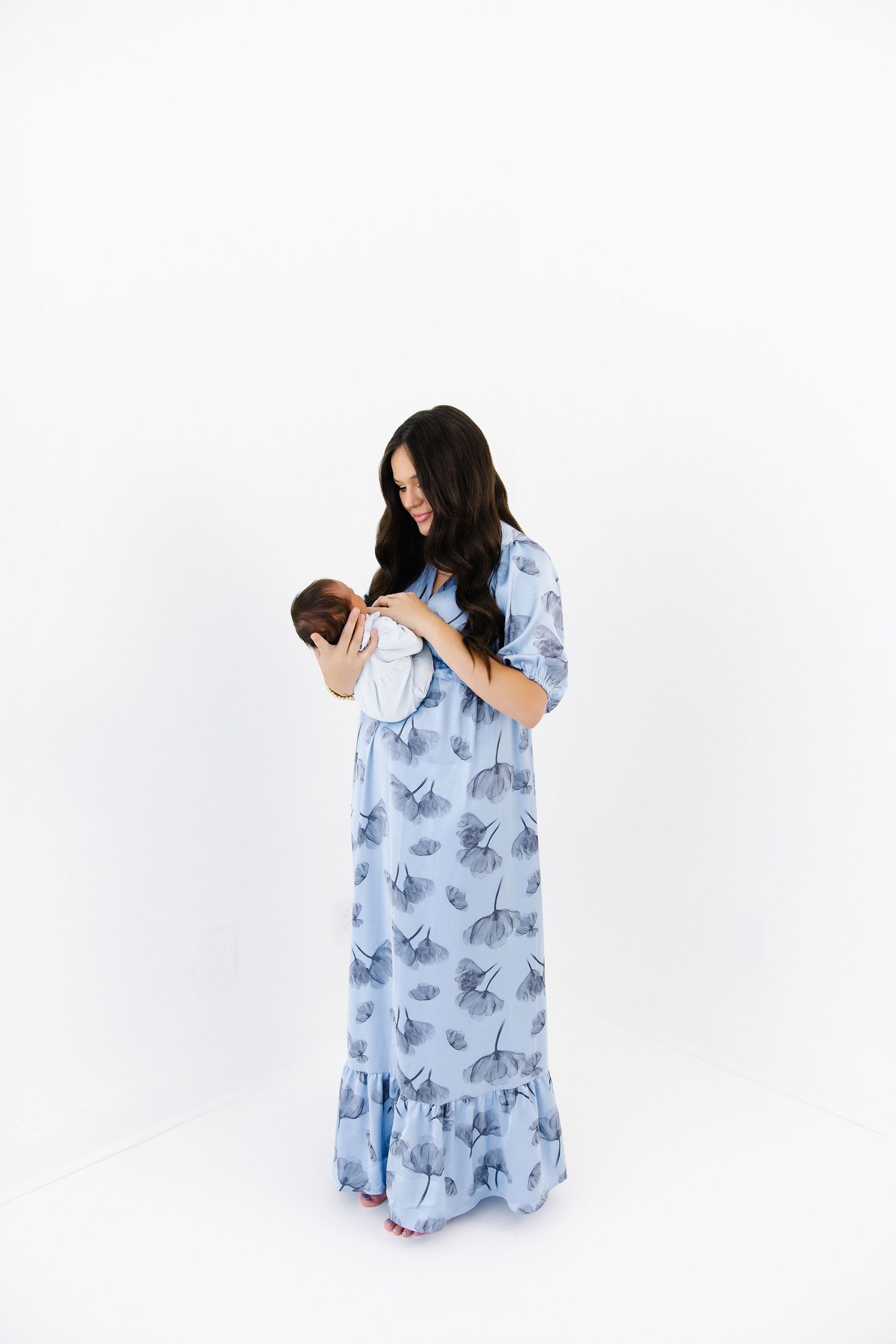 A woman wearing a blue floral dress holds a newborn baby in various tender poses against a white backdrop.