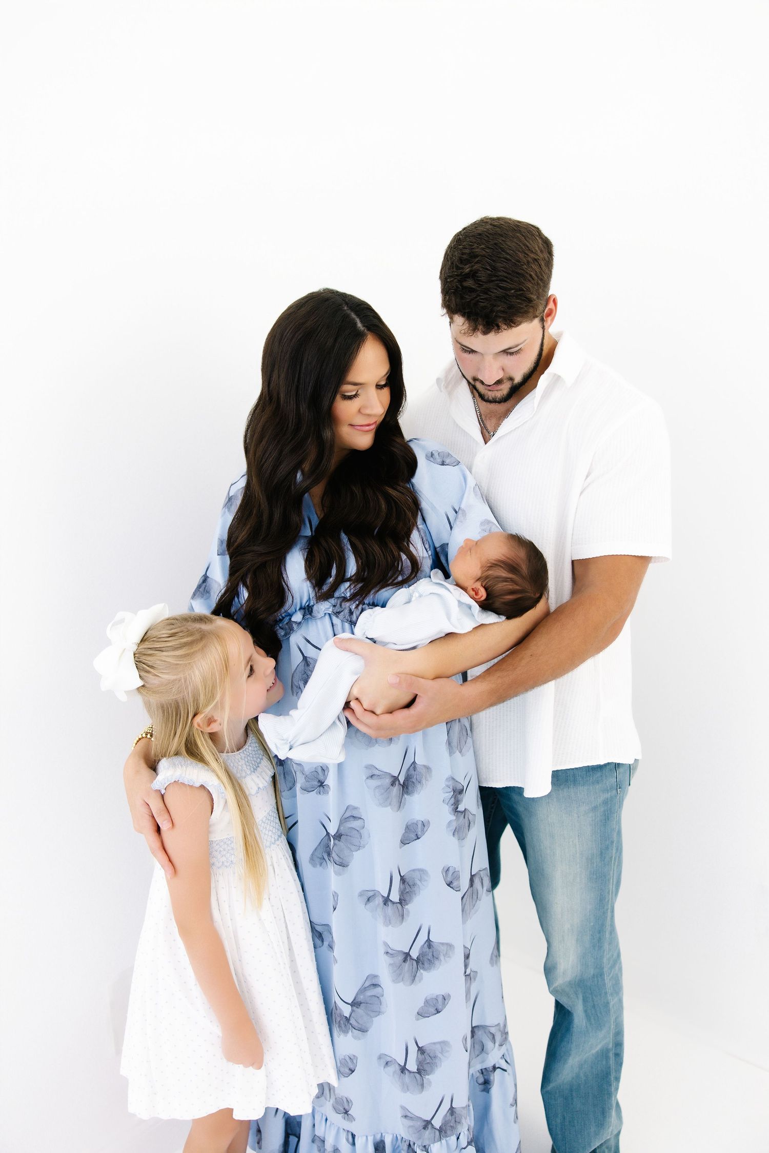 Family in coordinating blue and white outfits share tender moment against white studio backdrop.