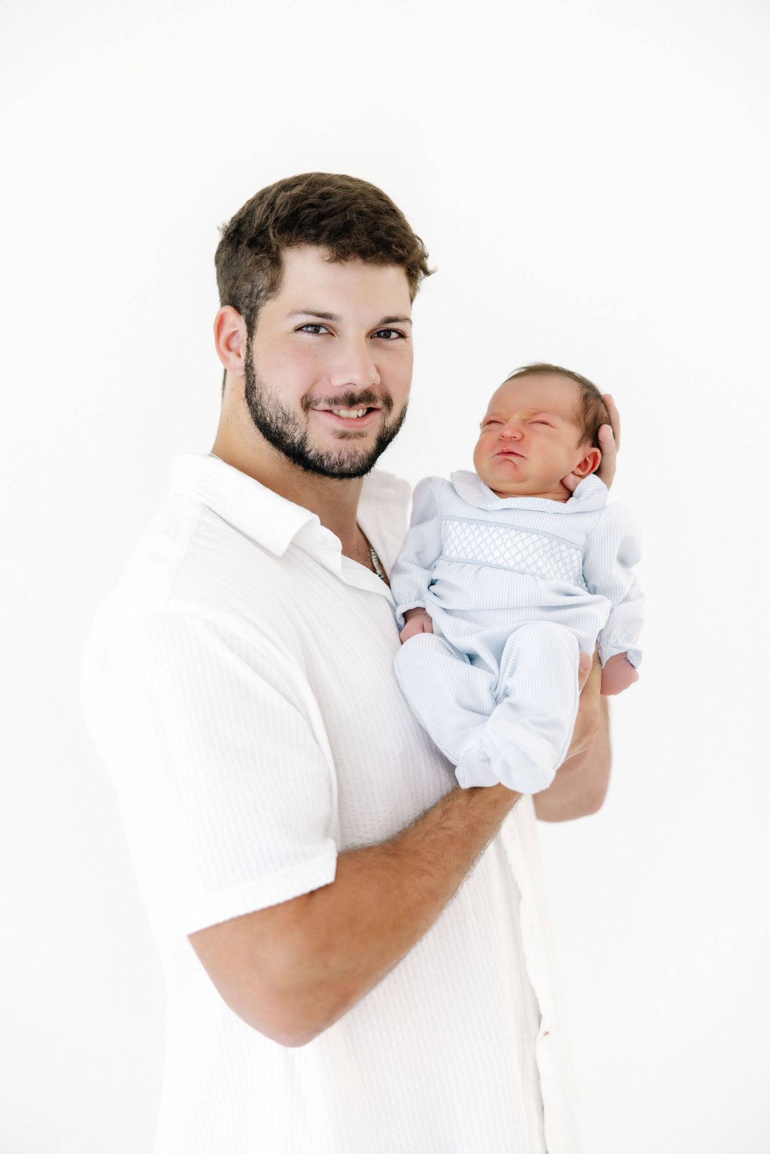 A man in a white shirt holds and smiles at a newborn baby dressed in white against a white background.