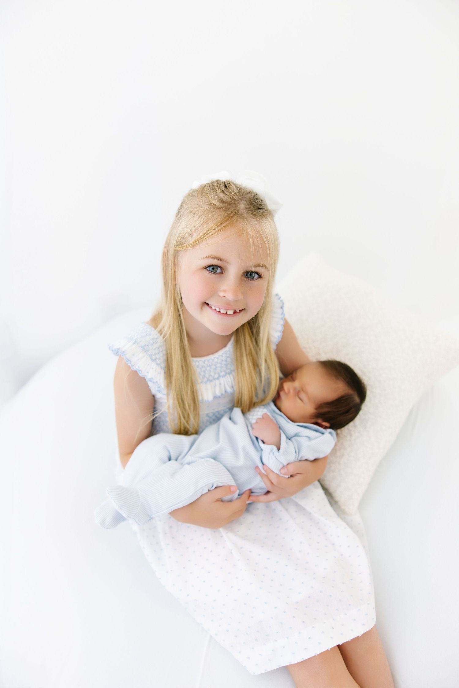 A young girl in a white dress holds a sleeping newborn baby while sitting on a white surface.